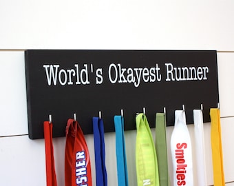 Könnte beinhalten: Ein rechteckiges, schwarzes Wandregal aus Holz mit dem weißen Schriftzug "World's Okayest Runner" auf der Vorderseite. Das Brett verfügt über acht silberne Metallhaken an der Unterkante, an denen jeweils ein farbiges Stoffband hängt.
