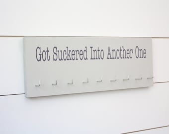 May include: A rectangular light gray wooden wall-mounted display rack featuring the dark gray text Got Suckered Into Another One printed on the front. The board includes ten evenly spaced silver metal hooks attached along the bottom edge.