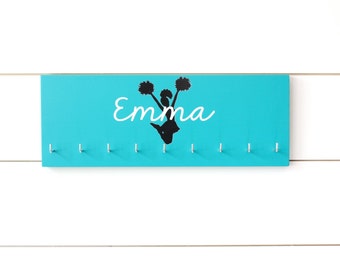 May include: A rectangular turquoise wall-mounted hook rack featuring the name Emma in white script lettering and a black silhouette of a cheerleader holding pom-poms. The wooden board includes nine evenly spaced silver metal hooks for hanging items.