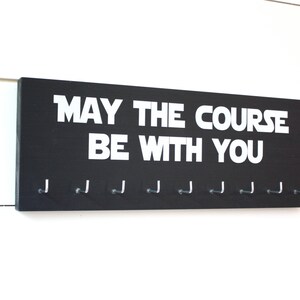 May include: Black wooden medal hanger with the text "May the course be with you" in white lettering. The hanger has 10 hooks for hanging medals.
