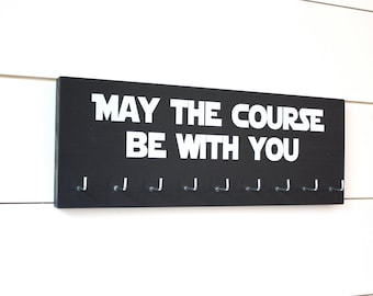 May include: A rectangular black wooden wall-mounted rack featuring the white text "MAY THE COURSE BE WITH YOU" printed in a bold, stylized font. The board includes nine evenly spaced silver metal hooks attached along the bottom edge.