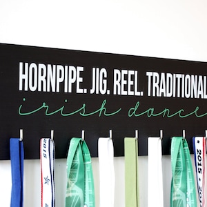 May include: Black wooden medal hanger with the text "Hornpipe. Jig. Reel. Traditional. Irish dance" in white and green lettering. The hanger has eight hooks for hanging medals.