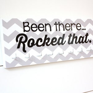 Peut inclure: Un porte-médailles en bois blanc avec un motif chevron gris et le texte "Been there...Rocked that." Le porte-médailles a 10 crochets argentés.