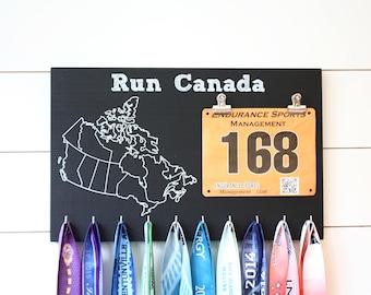 Könnte beinhalten: Schwarze Tafel mit einer weißen Umrandung von Kanada und dem Text "Run Canada". Eine Startnummer mit der Nummer "168" ist mit Clips an der Tafel befestigt. Acht bunte Medaillen hängen an Haken unter der Tafel.
