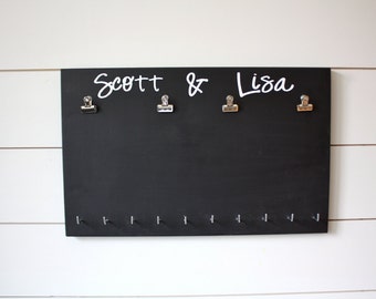 May include: A rectangular black wooden display board featuring the white text Scott & Lisa. The board includes four silver metal clips attached to the top section and a row of ten small silver hooks positioned along the bottom edge of the board.