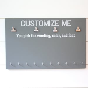May include: Grey wooden board with the text "CUSTOMIZE ME" and "You pick the wording, colour, and font". The board has four silver clips and 10 small silver hooks.