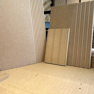 May include: A collection of light brown medium-density fibreboard panels featuring various surface textures. The panels include designs with vertical fluting, recessed grooves, and diamond patterns, displayed in a workshop setting.