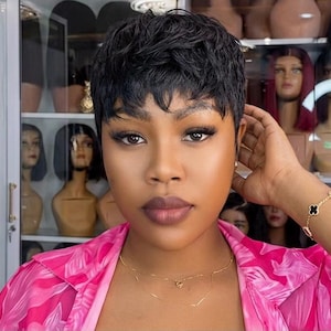 May include: A short, textured black pixie cut wig with layered strands. The hair is styled with a natural, piecey finish. The wig features a tapered back and sides with slightly longer, wispy bangs that frame the forehead, creating a voluminous look.
