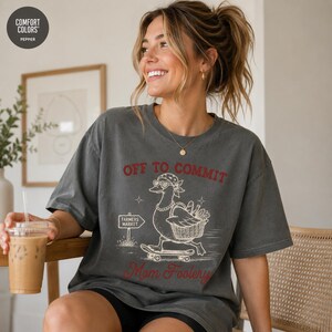 Può includere: Una t-shirt a maniche corte grigio antracite con la grafica di un'anatra che indossa una bandana, occhiali da sole a forma di cuore e una collana mentre va sullo skateboard. Il testo recita OFF TO COMMIT e Mom Foolery, con un cartello Farmers Market.