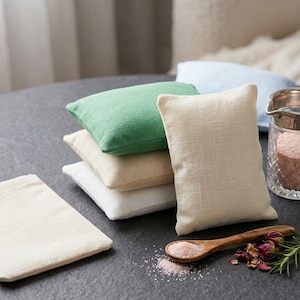 May include: A stack of small, rectangular linen pillows in various colors: green, beige, white, and light blue. A clear glass pitcher with pink salt, a wooden spoon with pink salt, and dried rose petals are also present. The items are arranged on a dark gray surface.