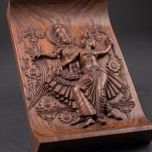 May include: A carved wooden decorative tray featuring a relief of two figures in traditional Indian attire. The figures are engaged in a dance, with intricate details in their clothing and surroundings. The wood has a rich, dark brown color.