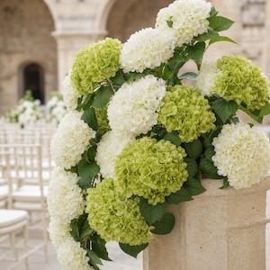 Puede incluir: Un gran arreglo floral con racimos de flores de hortensia blancas y verde lima con hojas de color verde oscuro, exhibido sobre un pedestal alto de piedra beige texturizada en un entorno al aire libre con filas de sillas blancas al fondo.