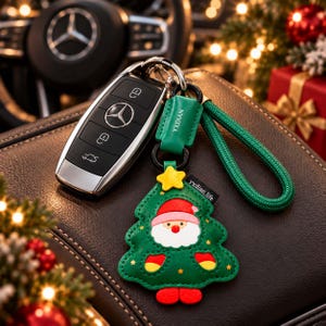 Könnte beinhalten: Ein schwarzer Mercedes-Benz Autoschlüsselanhänger an einem grünen Lederanhänger in Form eines Weihnachtsbaums mit Weihnachtsmann-Gesicht, einem gelben Stern an der Spitze und einer grünen geflochtenen Schlaufe. Mit YXDIAN-Prägung.