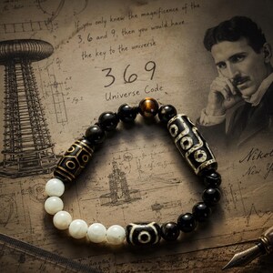 May include: A beaded bracelet featuring a mix of smooth white beads, black beads, and two cylindrical patterned beads with circular motifs. One bead displays a central eye design, while a single brown tiger eye bead sits at the top center.