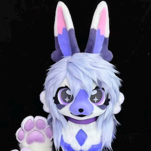 May include: A plush animal head mask featuring long, upright ears with pink and purple accents. The mask has shaggy light purple fur, large expressive eyes with star-shaped highlights, a purple nose, and a matching purple paw visible in the foreground.