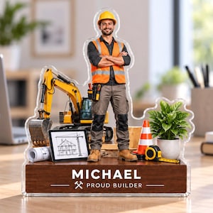 May include: A custom acrylic desk display featuring a construction worker in a yellow hard hat and orange safety vest, a yellow excavator, a traffic cone, a tape measure, a house blueprint, and a potted plant on a wood base labelled MICHAEL PROUD BUILDER.