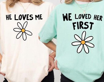 May include: Two short-sleeved t-shirts, one cream and one mint green, each featuring a daisy graphic and black text. The cream shirt reads HE LOVES ME, and the mint green shirt reads WE LOVED HER FIRST. Both shirts have a crew neckline.