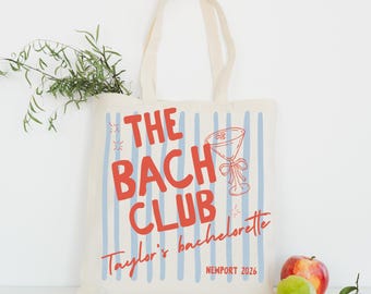 May include: A cream-colored canvas tote bag featuring vertical light blue stripes and red text that reads THE BACH CLUB, Taylor's bachelorette, and NEWPORT 2026. The design includes a red line-art illustration of a martini glass with a bow.