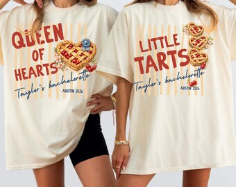 May include: Two off-white t-shirts featuring red and blue text and cherry pie graphics. The left shirt reads QUEEN OF HEARTS Taylor's bachelorette AUSTIN 2026. The right shirt reads LITTLE TARTS Taylor's bachelorette AUSTIN 2026 with three heart-shaped pies.
