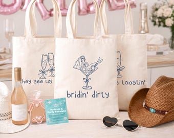 May include: Three off-white canvas tote bags with blue line art graphics. The center bag features a bride in a martini glass with the text 'bridin' dirty'. The side bags show champagne flutes and the text 'they see us' and 'toastin''.