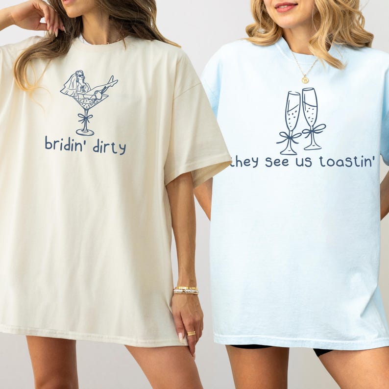 Może przedstawiać: Kremowy t-shirt z ciemnoniebieskim rysunkiem liniowym osoby w welonie w kieliszku do martini z napisem 'bridin' dirty' oraz jasnoniebieski t-shirt z dwoma kieliszkami do szampana i napisem 'they see us toastin'.