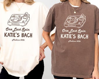 May include: Two short-sleeve crew neck t-shirts, one cream and one brown, each featuring a line art graphic of a record player. The text reads One Last Spin, KATIE'S BACH, and Madison 2026 in a dark brown font on the cream shirt and white on the brown.
