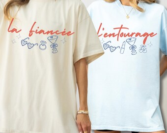May include: Two short-sleeve crewneck t-shirts. The cream shirt features red text reading la fiancée with blue line art of heart sunglasses, a diamond ring, and a martini glass. The light blue shirt features red text reading l'entourage with blue line art of heart sunglasses, lipstick, a martini glass, and cherries.