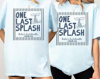 May include: A light blue t-shirt featuring a dark blue graphic with vertical stripes, a mermaid tail illustration, and the text "ONE LAST SPLASH", "Katie's bachelorette", and "CANCUN 2026" printed in a combination of block and script fonts.