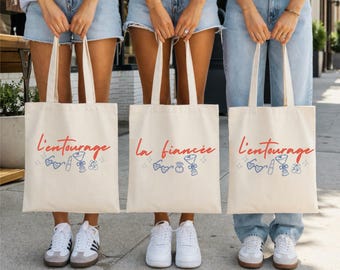 May include: Three off-white canvas tote bags featuring red cursive text and blue line art illustrations. Two bags display the text l'entourage with heart-shaped glasses, lipstick, a cocktail glass, and cherries. One bag displays la fiancée with glasses, a ring, and a cocktail glass.
