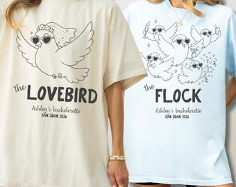 May include: A cream t-shirt featuring a black line drawing of a bird wearing heart-shaped sunglasses and a veil with the text 'the LOVEBIRD Ashley's bachelorette SAN JUAN 2026' and a light blue t-shirt with birds and 'the FLOCK Ashley's bachelorette SAN JUAN 2026'.
