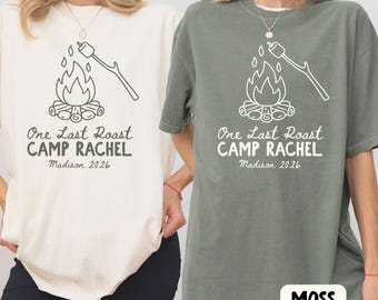 May include: Two short-sleeve crewneck t-shirts, one cream and one moss green, each featuring a dark graphic of a campfire with a marshmallow on a stick and the text One Last Roast CAMP RACHEL Madison 2026 printed in a casual font.