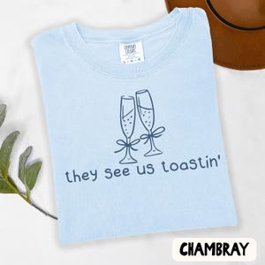 Może przedstawiać: Jasnoniebieski t-shirt z kr&oacute;tkim rękawem z ciemnoniebieską grafiką przedstawiającą dwa kieliszki do szampana przewiązane kokardkami oraz napis "they see us toastin'". Koszulka posiada metkę Comfort Colors przy dekolcie wskazującą rozmiar L.