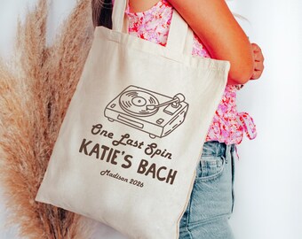 May include: A cream-colored canvas tote bag featuring a dark brown line art illustration of a record player. The text reads "One Last Spin", "KATIE'S BACH", and "Madison 2026" in a mix of script and bold sans-serif fonts printed on the front.