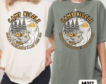 May include: Two short-sleeve t-shirts, one cream and one moss green, each featuring a circular graphic with a camping scene, a tent, a boat, and trees. The text reads CAMP NICOLE, NICOLE'S BACHELORETTE, and LAKE TAHOE 2026 in a bold, dark font.