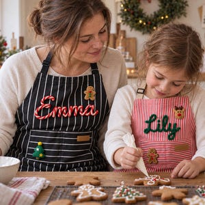 May include: Two aprons featuring vertical stripes and personalised names in raised lettering. The black apron displays the name Emma with a gingerbread man patch, while the red apron displays the name Lily with a reindeer patch.