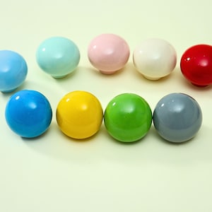 May include: A collection of nine spherical cabinet knobs in a variety of solid colors including light blue, mint green, soft pink, cream, bright red, deep blue, yellow, lime green, and gray, each featuring a smooth, glossy, reflective finish.