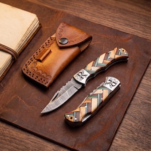 May include: Two folding pocket knives with Damascus steel blades and handles featuring a multi-colored herringbone wood pattern. Each knife has a metal bolster engraved with the letters DK. A tan leather sheath with decorative stitching is included.