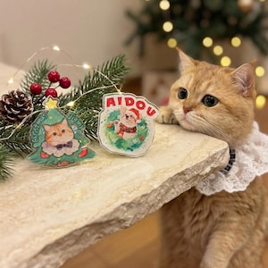 May include: A golden cat with a white lace collar gazes at the camera. Two Christmas-themed acrylic ornaments are on a table. One is a Christmas tree with a cat illustration, the other has a dog illustration and the word "AIDOU".