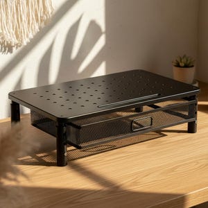 May include: Black metal monitor stand with a perforated top and a mesh drawer. The stand is on a wooden desk, with a small potted plant and a mug in the background. The stand is designed to elevate a computer monitor.