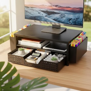 May include: Black desk organizer with a monitor on top. The organizer has a drawer, shelves, and side compartments filled with pens, pencils, notebooks, and a calculator. A small succulent plant is in the drawer. The background shows a window and a landscape painting.