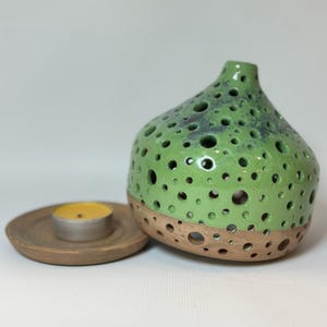 May include: A ceramic candle holder with a rounded, bulbous shape featuring numerous circular cutouts. The upper portion is glazed in a vibrant lime green with dark speckles, while the base remains unglazed brown clay. A small yellow tea light sits on a matching brown ceramic saucer.