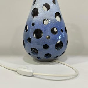 May include: A pear-shaped ceramic lamp with a blue and white gradient glaze and circular cutouts. It measures 24 cm in height and 17 cm in width. The lamp includes a white power cord with an inline switch for operation.