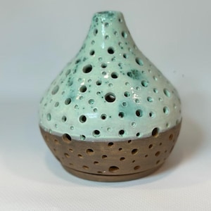May include: A ceramic vase with a tapered neck and a rounded base, featuring a light mint green glazed upper section and a raw brown clay lower section. The surface is covered in circular cutouts of varying sizes. Width: 12 cm, Height: 13.5 cm.