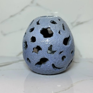 Puede incluir: Un recipiente esférico de cerámica azul claro con una superficie texturizada y craterizada, con múltiples aberturas irregulares. El objeto mide 16 cm de altura y 16 cm de ancho, presentando un interior hueco y un acabado suave.