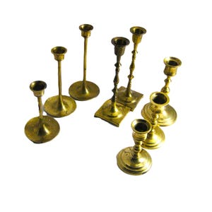 May include: A collection of vintage brass candlesticks in various shapes and sizes. The candlesticks have round and square bases, with slender stems and candle holders. The brass has a warm, aged patina, and the candlesticks are arranged on a white background.