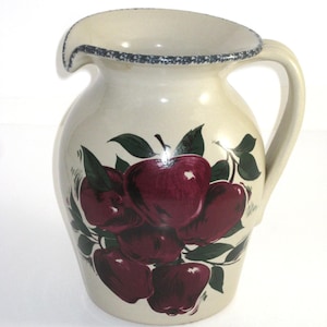 May include: Cream-colored ceramic pitcher with a handle and spout. The pitcher is decorated with a cluster of red apples and green leaves. The rim has a dark blue speckled design. A decorative kitchenware item.