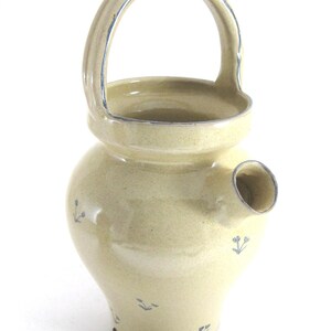 May include: A handmade, light yellow ceramic pitcher with a handle and a spout. The pitcher has a rounded body and is decorated with small blue floral designs. The handle and rim are accented with a thin blue line.