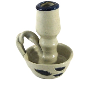 May include: A handmade ceramic candlestick holder in a light beige color with a dark blue rim. The holder has a handle and a small bowl with leaf-shaped cutouts. The candlestick is a unique piece of home decor.