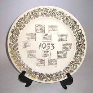 May include: A vintage cream-colored plate featuring a calendar for the year 1953. The plate has a gold floral border and individual month calendars in black. The year "1953" is prominently displayed in the center.