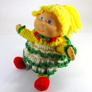 May include: A vintage doll with yellow yarn hair styled in a ponytail. The doll wears a crocheted dress with yellow, white, green, and red accents. Red crocheted shoes complete the outfit. The doll has blue eyes and a smiling expression.
